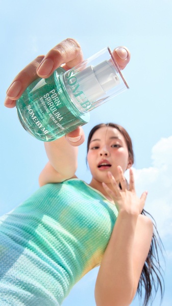 A close-up, low-angle shot of a woman holding a spirulina repair serum bottle against a bright, clear blue sky background.